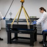 Two lab technicians use lifting slings and a hoist to safely move a granite surface plate in a metrology lab.