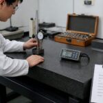 A metrology technician calibrates a granite surface plate using precision measuring tools in a quality control lab.