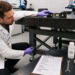 A male metrology technician tightens a leveling foot on a surface plate stand while checking a bubble level on a granite surface plate in a calibration lab.