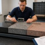 A metrology technician measures flatness on black, gray, and pink granite surface plates in a calibration lab.